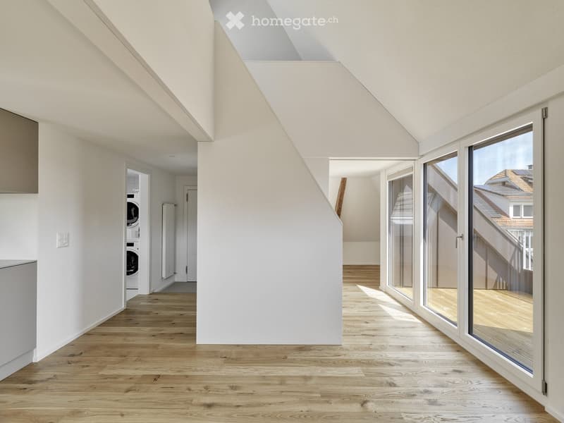 Bright, open loft-style room with wooden flooring, white walls, and large windows offering a view of the roof. A private washing machine is visible in an adjacent area.