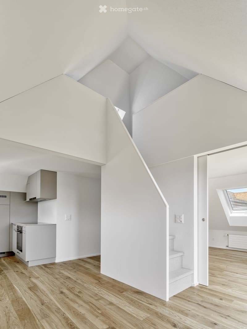 Bright, open loft-style space with wooden flooring, white walls, and a staircase leading to an upper level. The kitchen area features white cabinets and stainless steel appliances, while the upper level includes a skylight window.