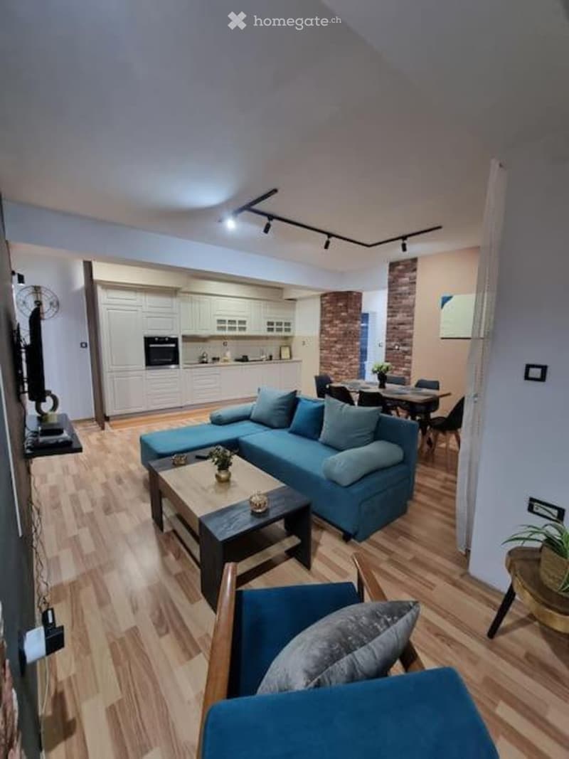 Modern living room with a blue sectional sofa, coffee table, dining area with chairs and table, kitchen with white cabinets and an oven, and a fan near the TV.