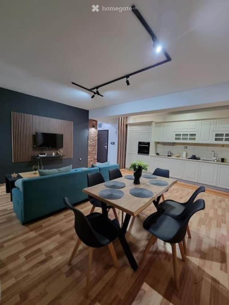 A modern, open-plan living and dining area with a blue sofa, a wooden dining table with black chairs, a television mounted on a dark wall, and a white kitchen with cabinets, an oven, and a sink.