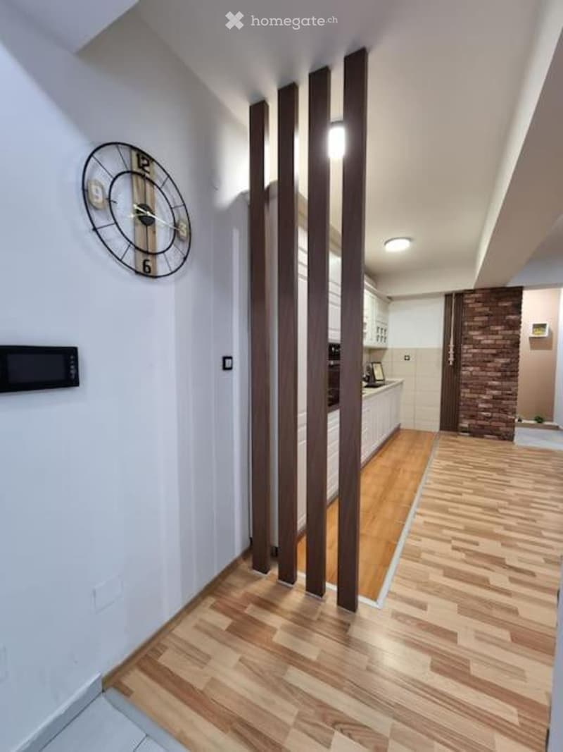 Modern hallway with light wooden flooring, white walls, and a decorative clock on the wall. The hallway leads to a kitchen area with white cabinets and a brick accent wall.