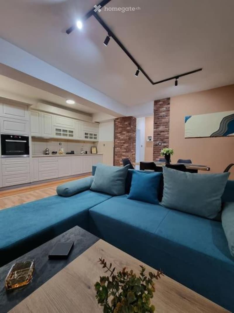 Open-plan living and dining area with a modern blue sectional sofa, wooden coffee table, sleek white kitchen cabinets, and a dining table with chairs. The space features recessed lighting and a decorative brick accent wall.
