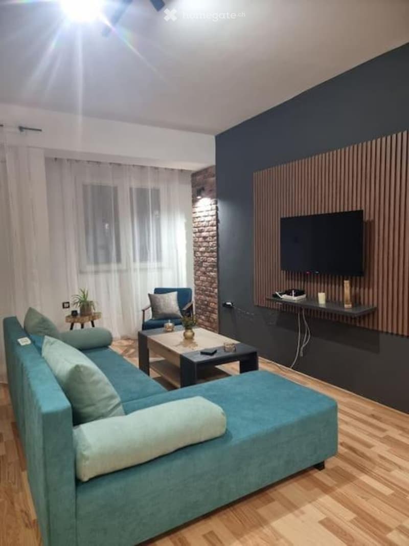 Modern living room with a blue sectional sofa, coffee table, armchair, small side table with a plant, and a TV mounted on a dark accent wall. The space features exposed brick, wooden flooring, and large windows with white curtains.