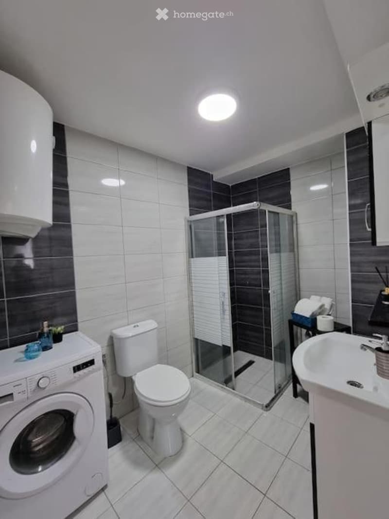 Modern bathroom with white tiles, a toilet, sink, shower, washing machine, and a small shelf with towels.