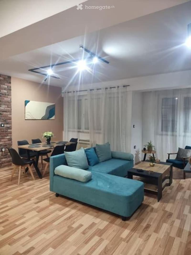 Modern living and dining area with a blue sectional sofa, coffee table, dining table with chairs, and decorative elements including a flower vase and a mirror on the wall.