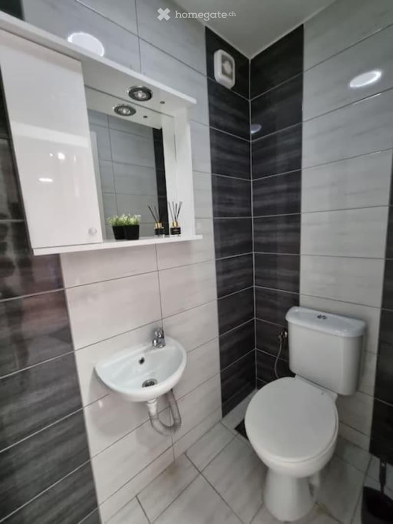 Modern bathroom with white tiles, dark grey accent wall, white sink, toilet, and mirrored cabinet.