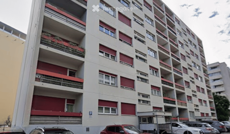 Multi-story apartment building with red shutters, balconies, and a parking area.