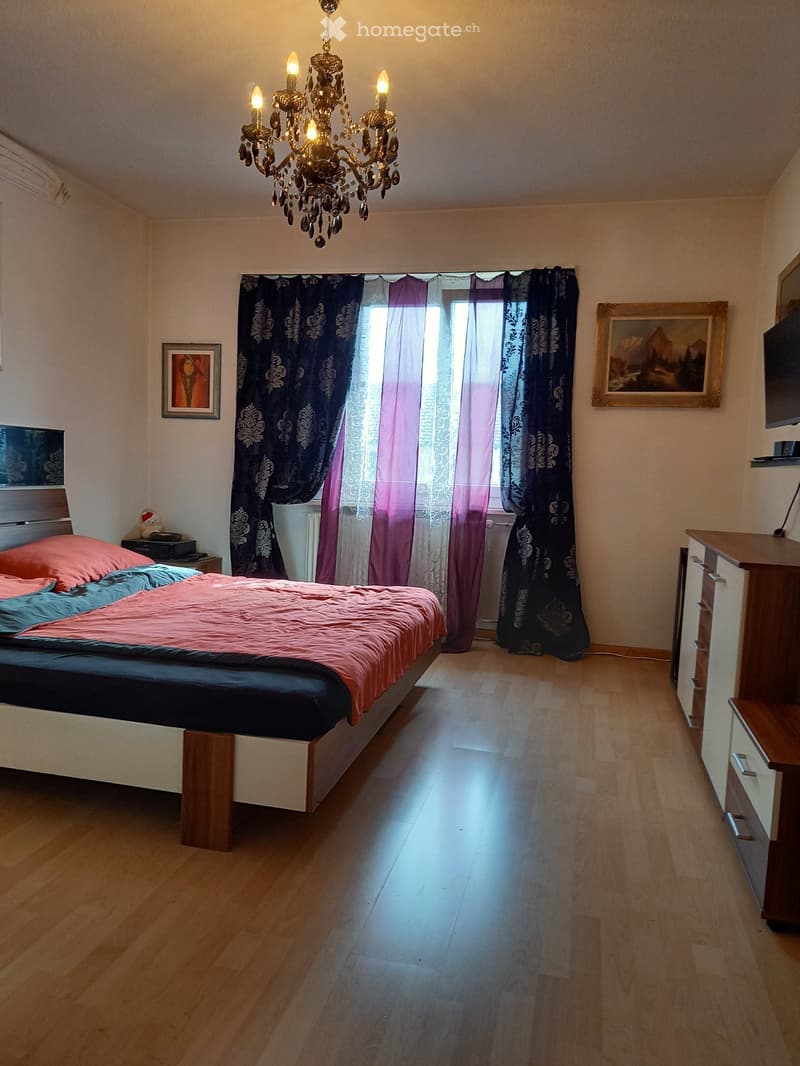 Bedroom with wooden flooring, a bed with pink and blue bedding, a chandelier, nightstand, and a cabinet.