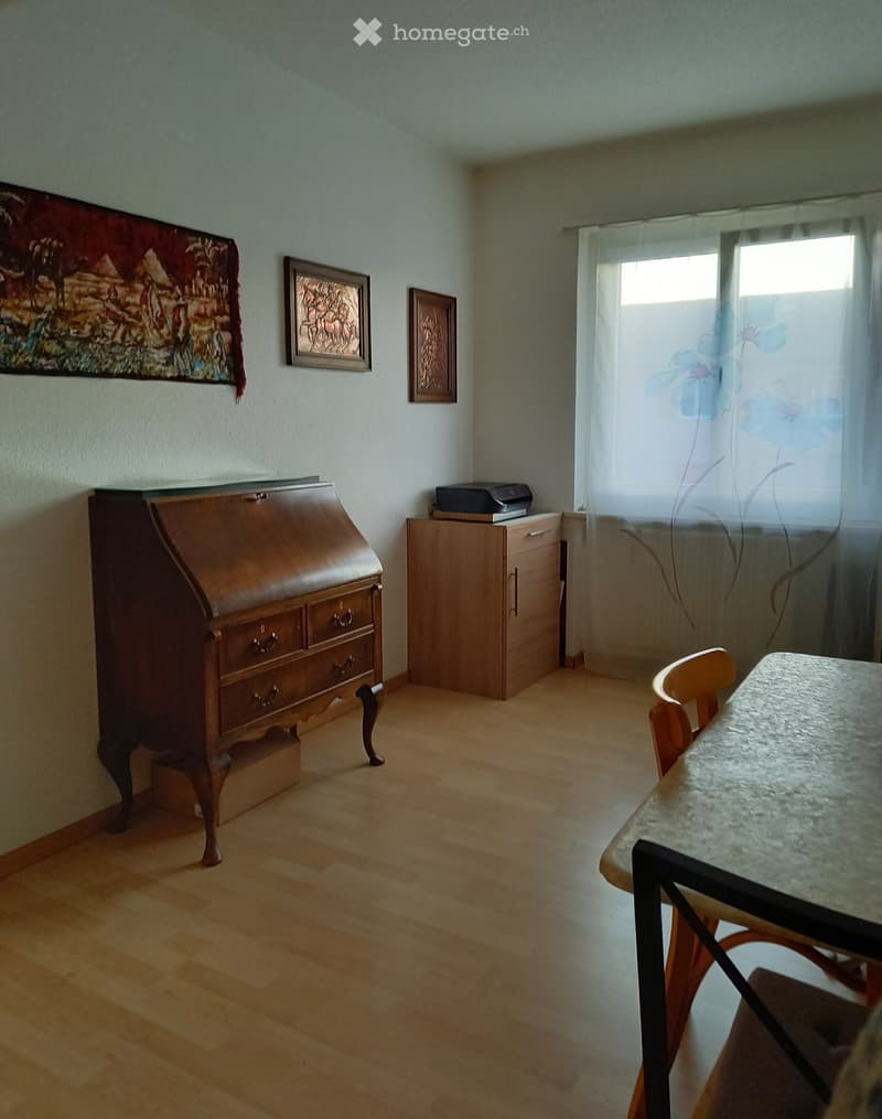 A small, well-lit room with wooden flooring, a wooden desk, a chair, a small table with a marble top, a cabinet, and a printer on top of the cabinet. The walls are adorned with framed artwork and a tapestry.
