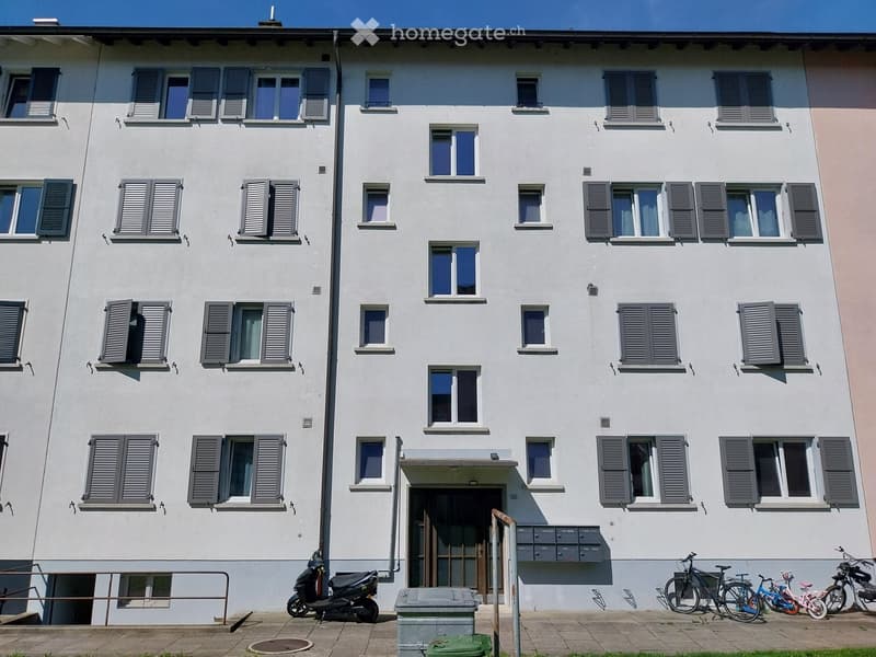 Four-story residential building with white facade, multiple windows with shutters, and a small entrance area with bicycles and a scooter parked nearby.