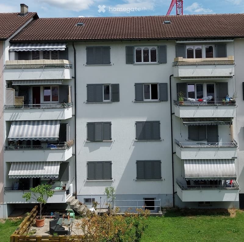 A three-story white apartment building with multiple balconies, grey shutters, and a small garden area in front.