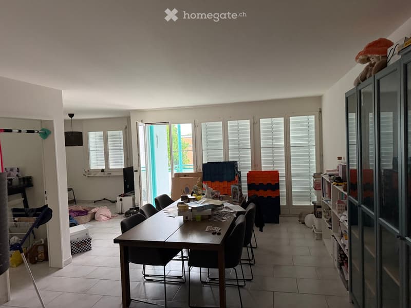 Bright room with large windows and shutters, a wooden table surrounded by chairs, a TV screen, and a cabinet with shelves.