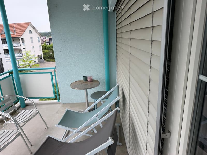 Balcony with a small round table, three chairs, and a view of neighboring buildings and greenery.