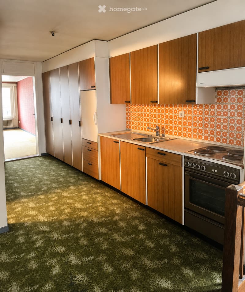 Kitchen with wooden cabinets, a sink, stove, oven, and refrigerator. The backsplash features a retro orange and white floral tile design, and the floor has a green patterned carpet.