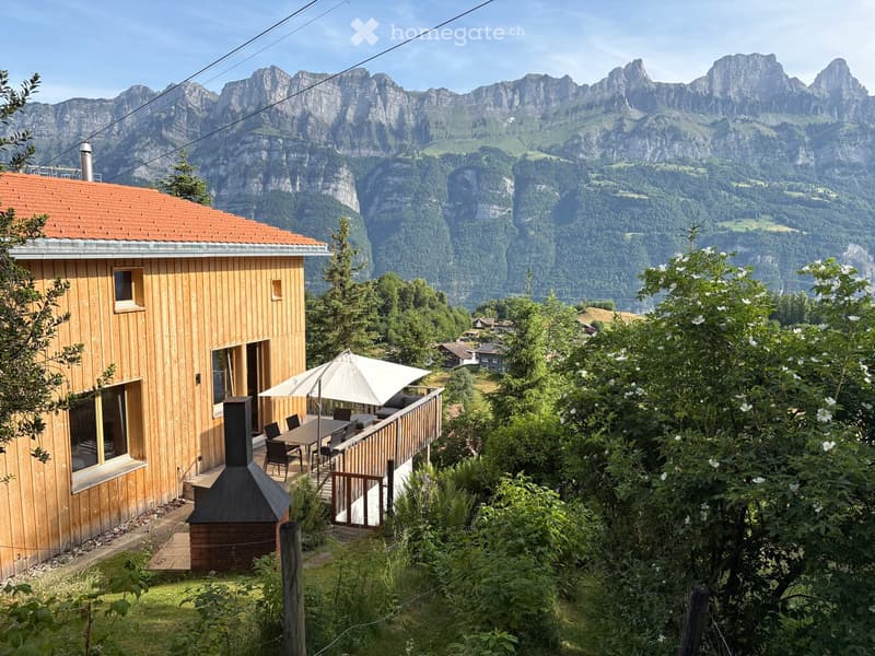 A wooden house with a red-tiled roof, large windows, and a balcony overlooking a scenic mountain range. The balcony features a table, chairs, and an umbrella, surrounded by lush greenery.