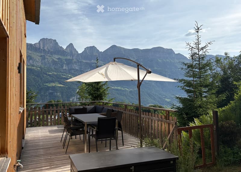 Balcony with wooden decking, outdoor furniture including a table, chairs, and a sofa, umbrella, and scenic mountain view.