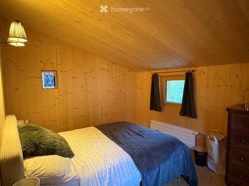 Cozy wooden bedroom with a double bed, nightstand, lamp, and a framed picture on the wall. The room features wooden walls and a slanted ceiling, with a window covered by dark curtains.