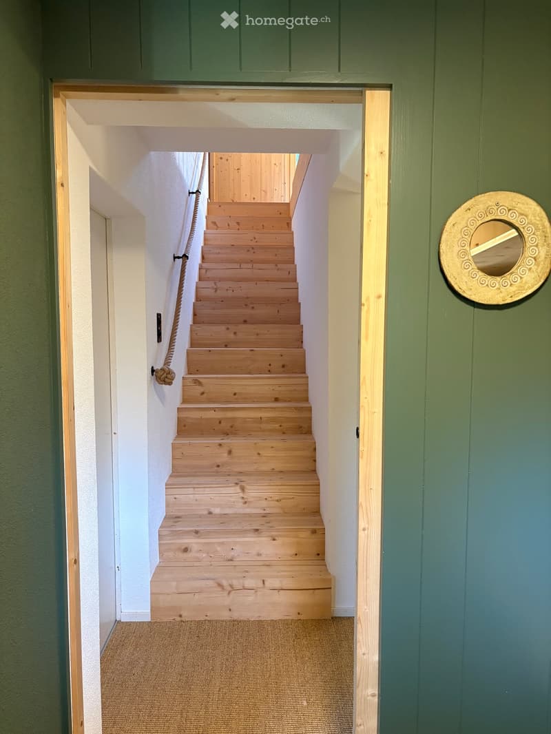 A narrow hallway with a wooden staircase leading upwards, featuring a rope handrail and a decorative round mirror on the wall.