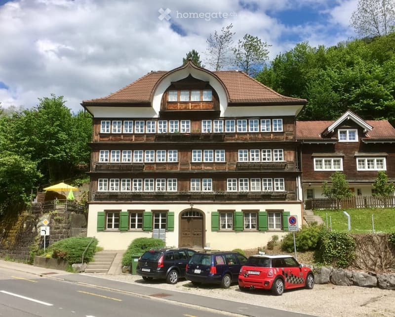 A three-story traditional house with a wooden facade and green shutters, featuring multiple windows and a central entrance. The building is surrounded by greenery and has several parked cars in front.