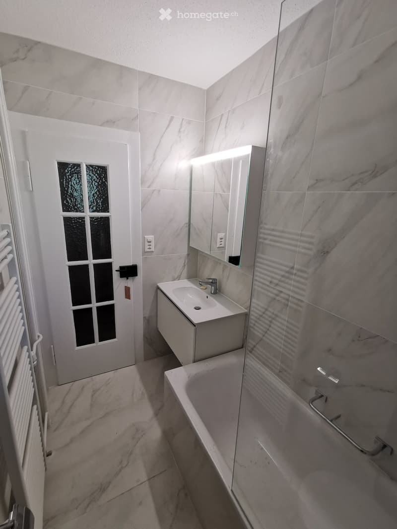 Modern bathroom with marble tiles, a white sink, bathtub, and a large mirror with built-in lighting. Features a frosted glass window and a radiator.