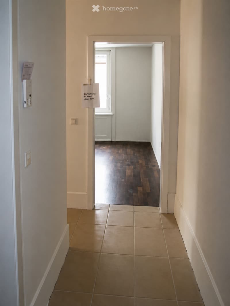 Empty hallway with tiled floor and wooden flooring visible in the adjacent room. Light-colored walls and a white door frame lead to another space.
