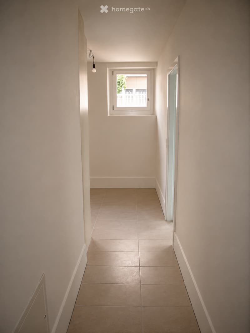 A narrow hallway with beige tiled flooring and light-colored walls. Features a single window with white frame and a small light fixture on the wall.