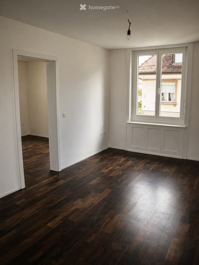 Empty room with dark wooden flooring, white walls, and large windows showing a view of neighboring buildings.