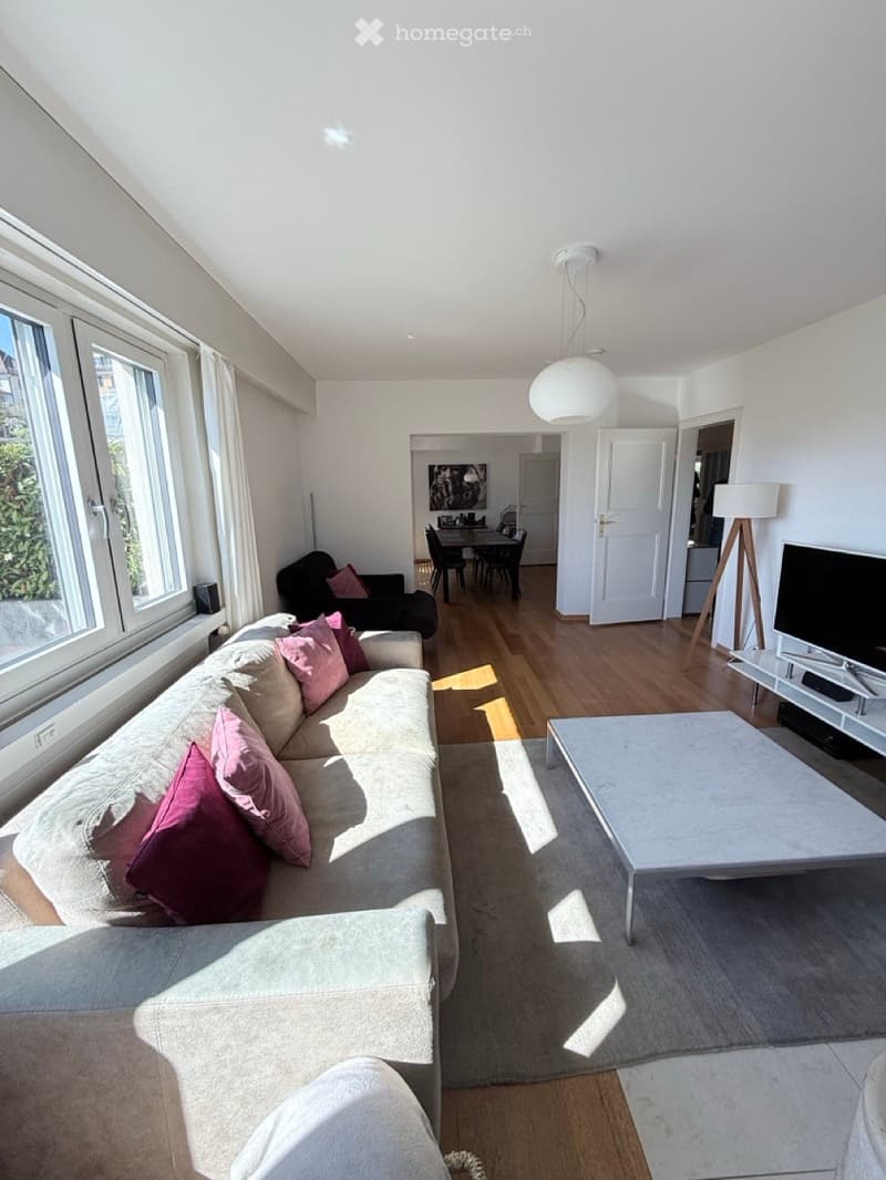 Bright living room with large windows, a beige sofa with pink cushions, a coffee table, an armchair, a dining area with a table and chairs, and a TV on a stand.