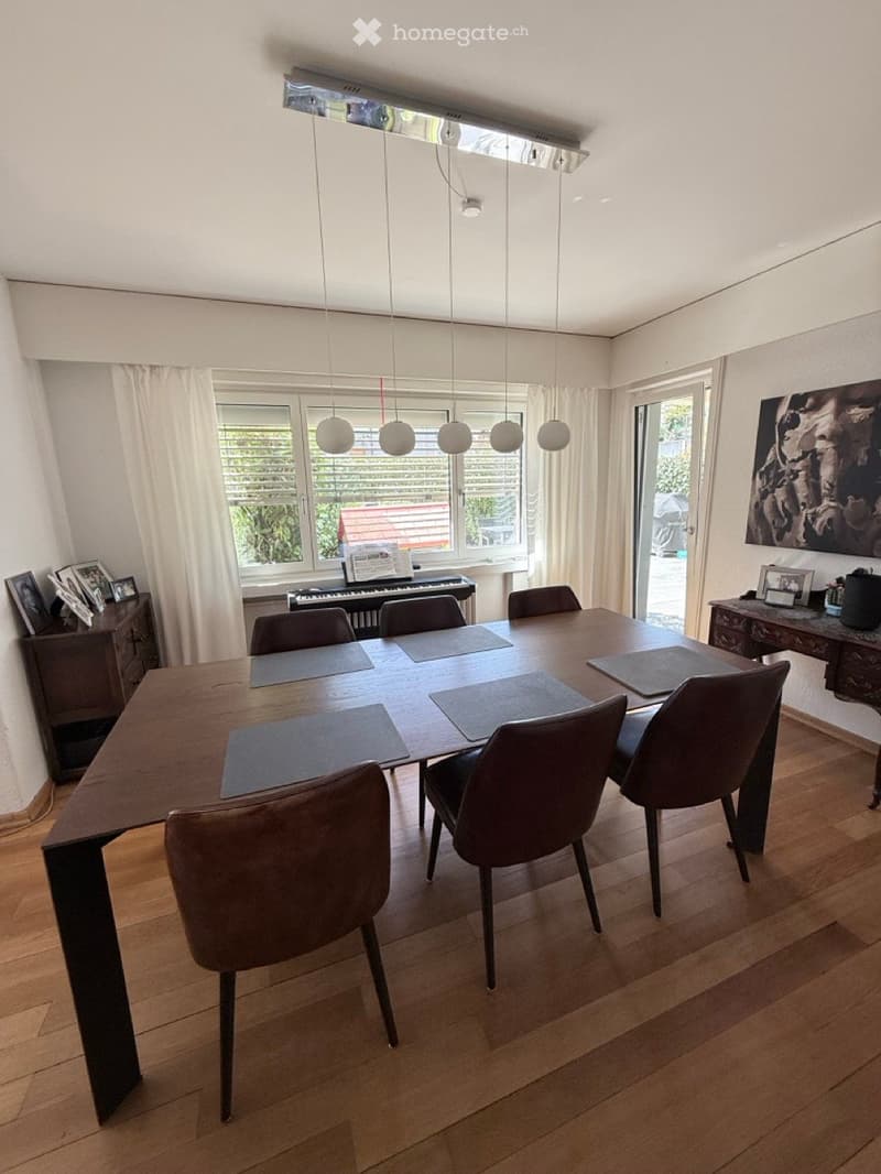 Dining room with a wooden dining table, six chairs, a piano, and large windows with curtains. The room features a modern pendant light fixture and a small cabinet with picture frames on top.