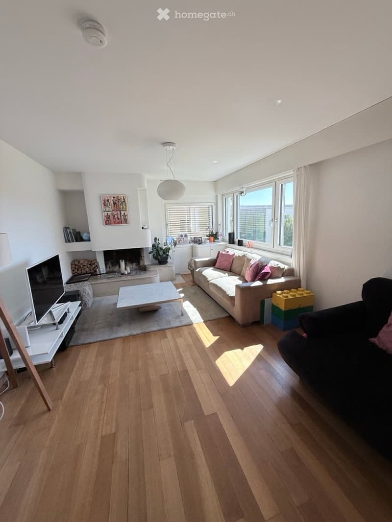 Bright living room with wooden flooring, a fireplace, a sofa, coffee table, TV stand, and a potted plant. Large windows allow natural light to fill the space.