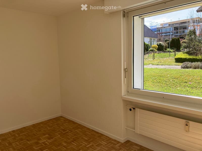 Empty room with wooden flooring, white walls, and a large window showing a green outdoor area with grass and trees.