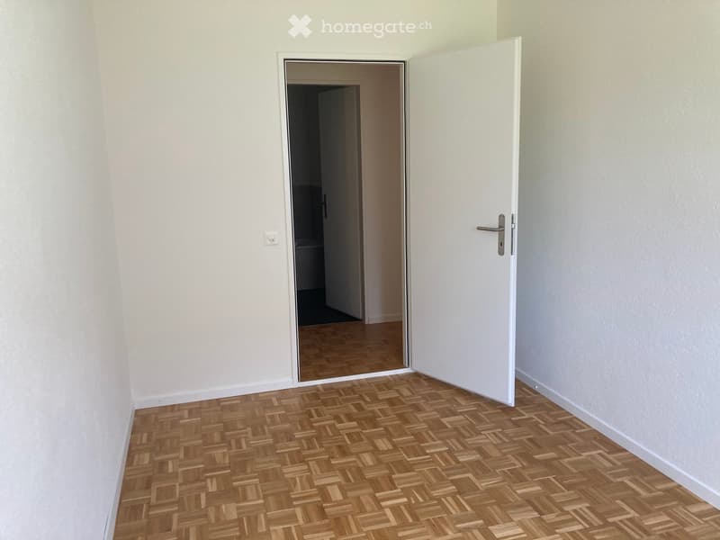 Empty room with light-colored walls and wooden herringbone flooring, featuring an open door leading to another room.