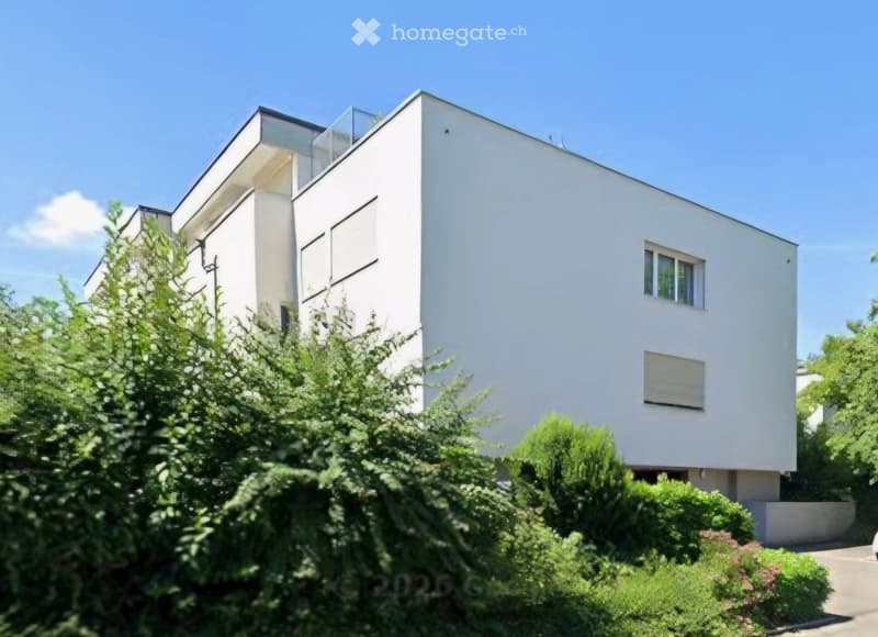 Modern two-story white house with large windows and a balcony on the upper level, surrounded by greenery.