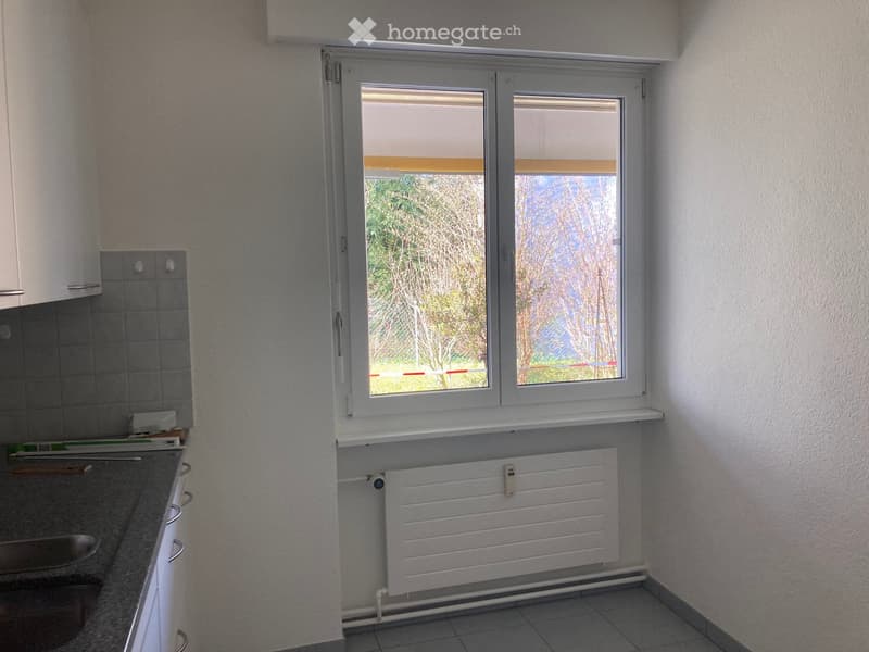 Empty kitchen with white walls, tiled floor, window with blinds, radiator, and countertop.