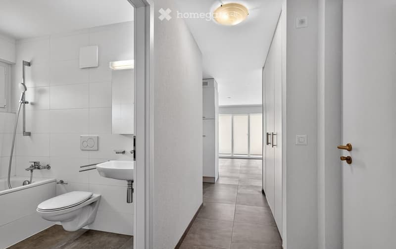 Modern bathroom with white tiles, bathtub, toilet, sink, and shower. The hallway leads to another room with large windows.