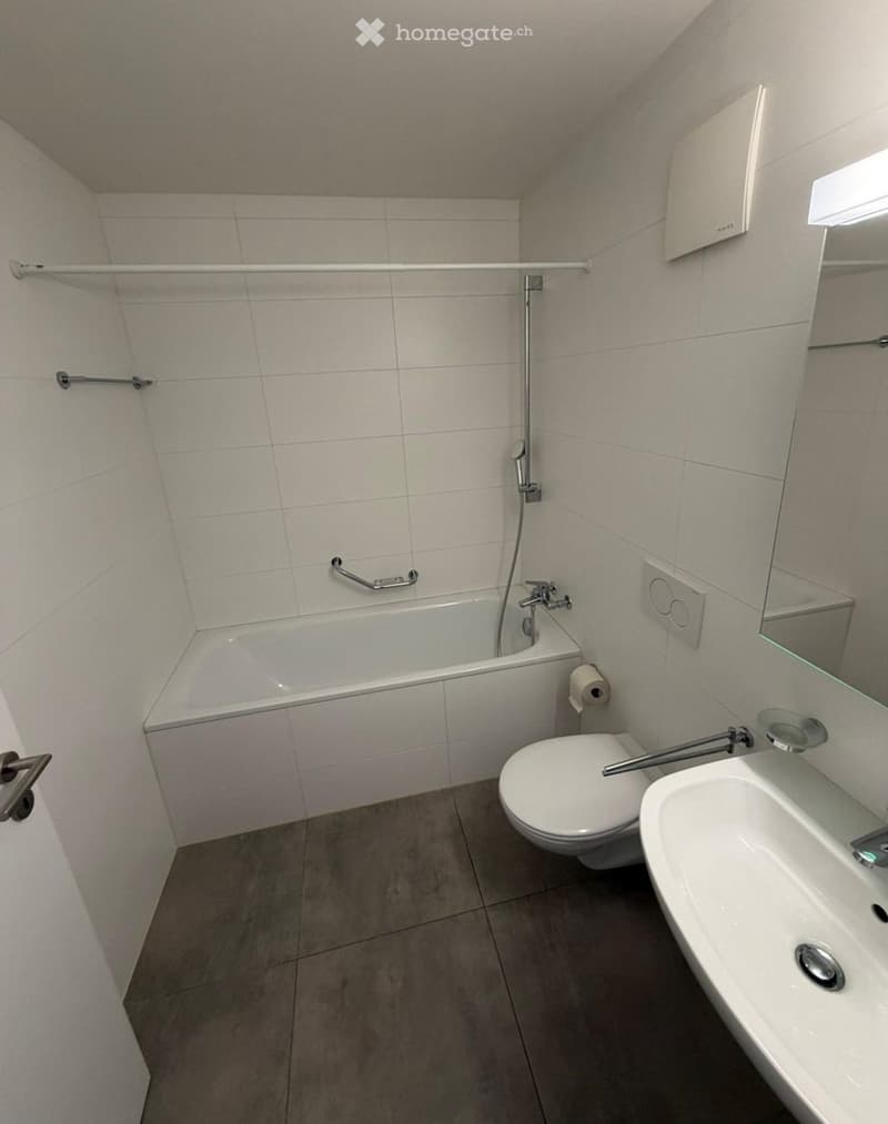 Modern bathroom with a bathtub, toilet, sink, and mirror. Features tiled walls and a grey tiled floor.