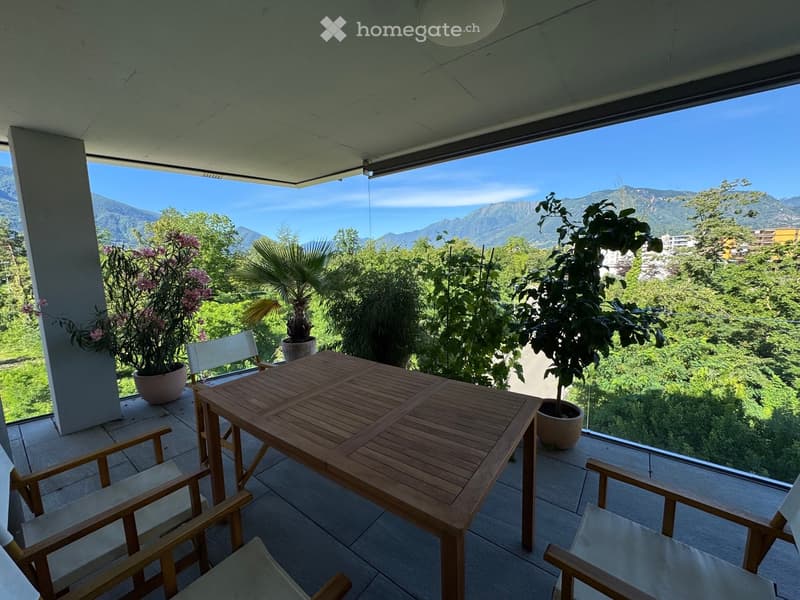 Balcony with wooden table and chairs, potted plants, and scenic mountain view.