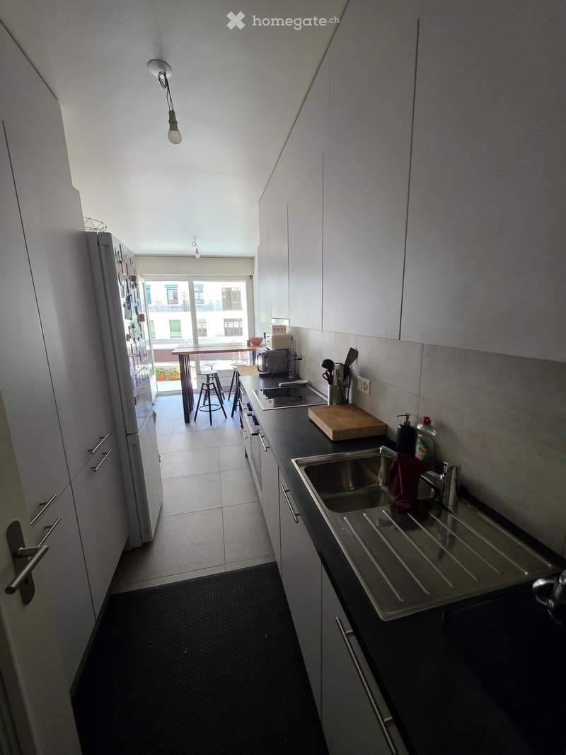 Modern kitchen with white cabinets, stainless steel sink, and black countertops. Includes a refrigerator, cutting board, and a small dining area with stools visible through an open doorway.