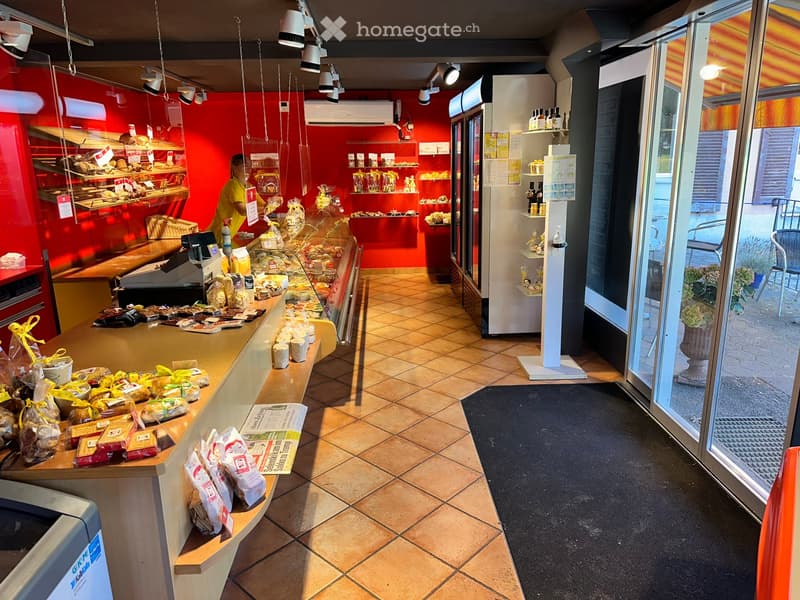 Bakery interior with red walls, tiled floor, counter displaying bread and pastries, shelves with products, and a refrigerator.