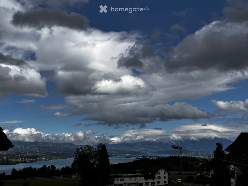 Panoramic view of a lake surrounded by mountains under a partly cloudy sky, with a few buildings and trees in the foreground.