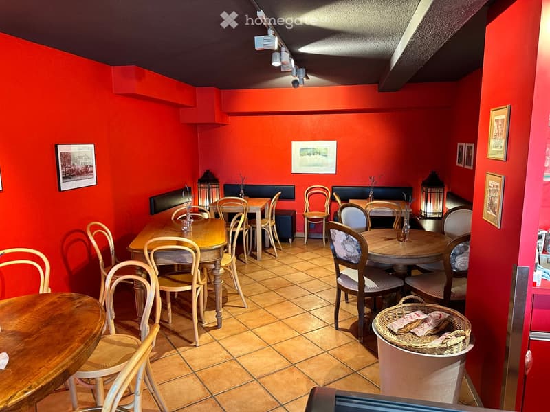 A cozy restaurant with red walls, wooden tables, and chairs arranged around the space. Lanterns and picture frames adorn the walls, and a basket with menus sits on a counter.