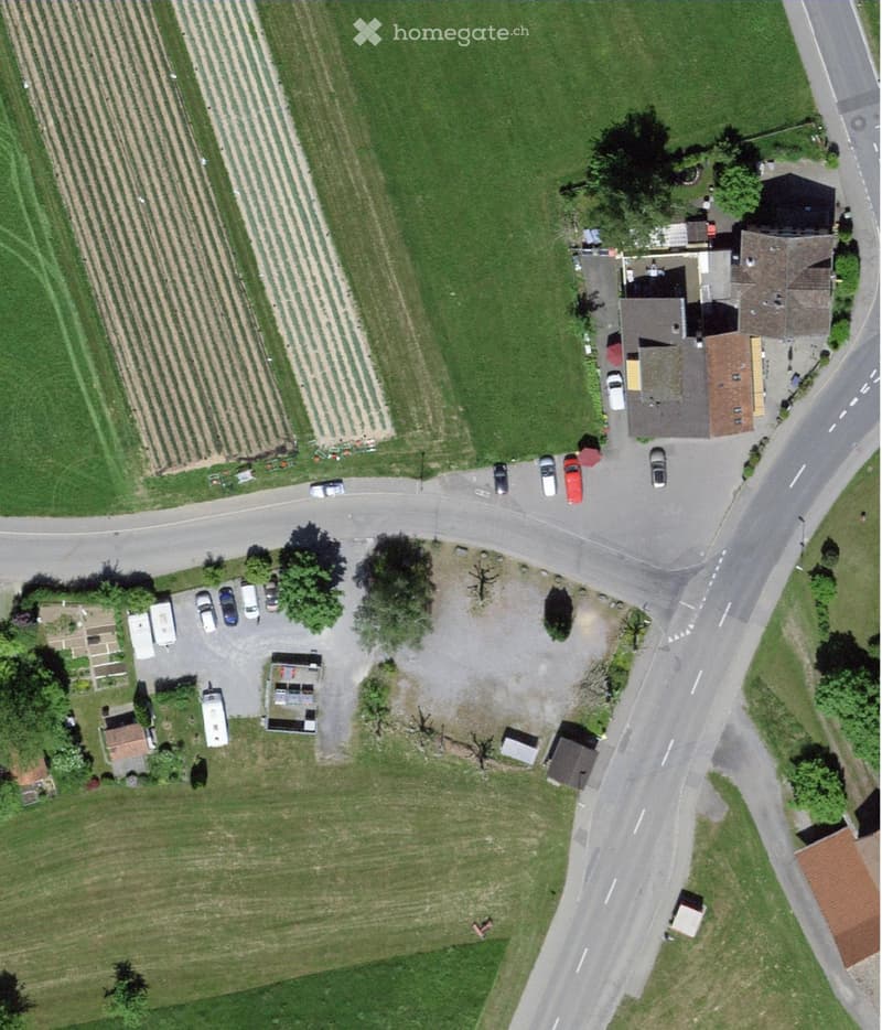 Aerial view of a residential building with a driveway, parked cars, and surrounding greenery, including fields and trees.