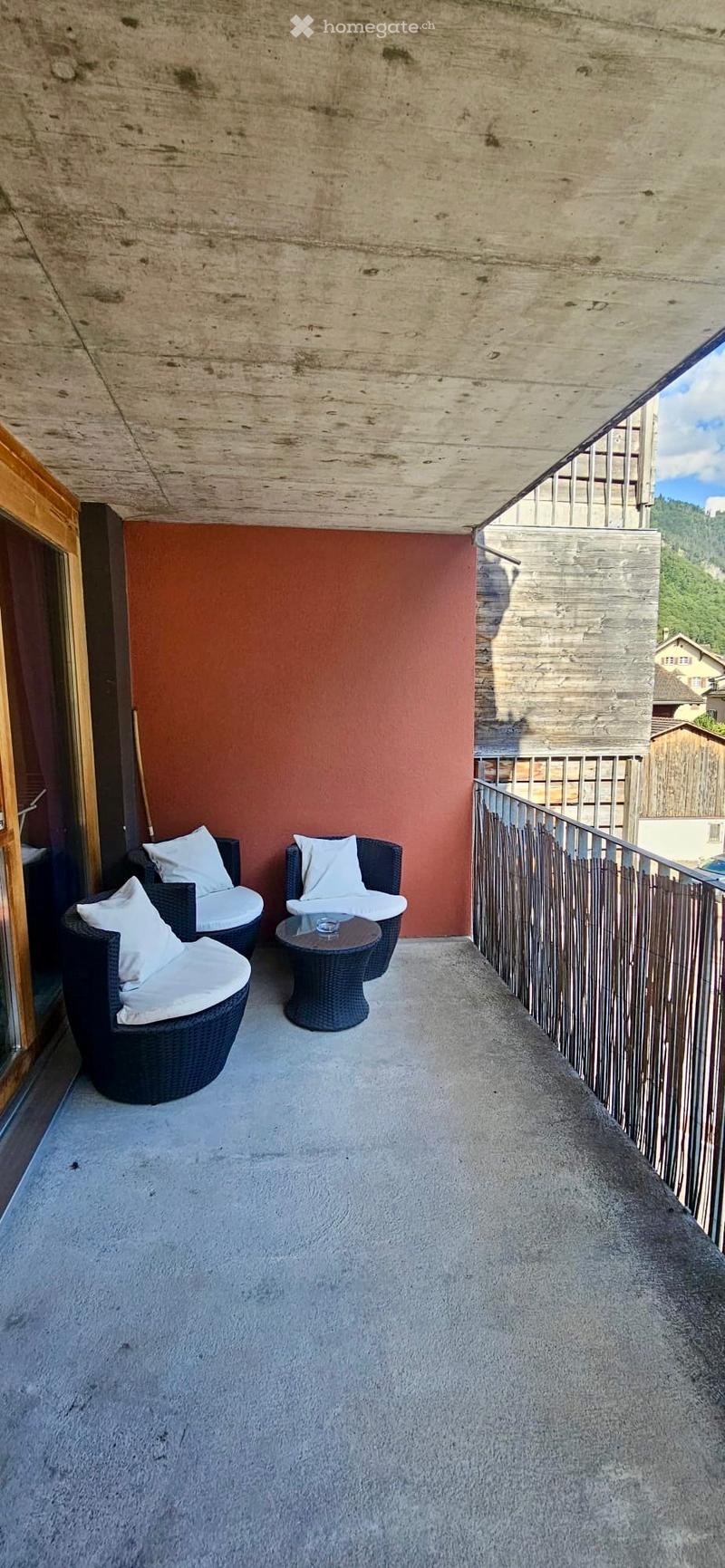 Balcony with a red accent wall, two wicker chairs with white cushions, a small round table, and a metal railing overlooking a scenic view.