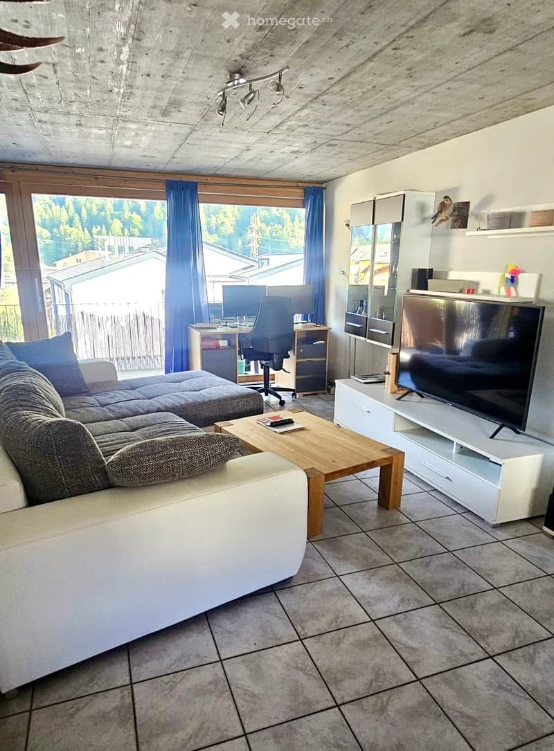 Modern living room with a grey sectional sofa, wooden coffee table, flat-screen TV on a white stand, desk with chair, and large windows with blue curtains.