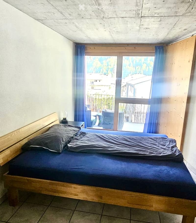 A modern bedroom with a wooden bed, nightstand, and lamp, featuring large windows with blue curtains that offer a view of the outdoor area.