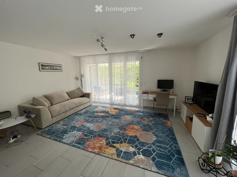Bright living room with a beige sofa, colorful geometric rug, coffee table, desk with chair, television, and floor lamp. Large windows with sheer curtains let in natural light.