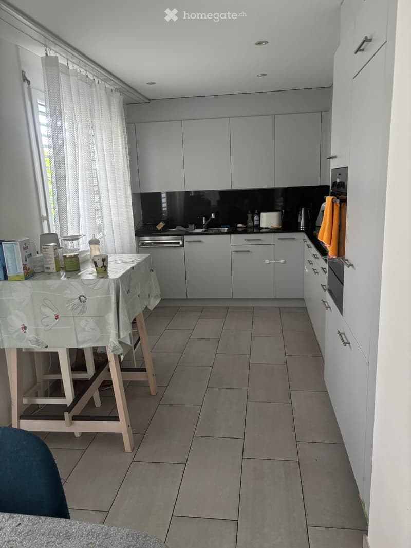 Modern kitchen with white cabinets, tiled floor, and a small table with a floral tablecloth. Visible items include a cup, a bottle, and a dish rack.