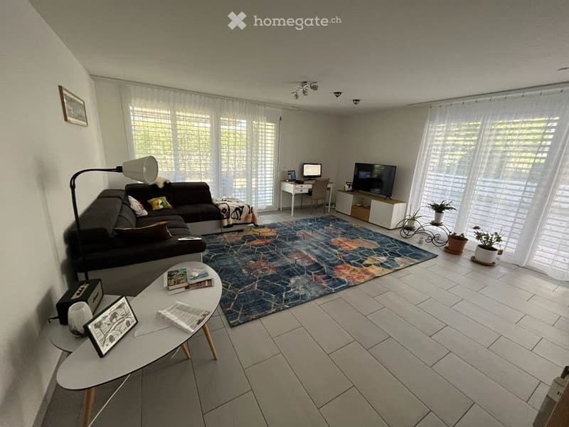 Modern living room with white tiled flooring, large windows with blinds, a dark grey sectional sofa, a colorful patterned rug, a white coffee table with magazines, a floor lamp, a TV stand with a flat-screen TV, a small desk with a laptop, and potted plan