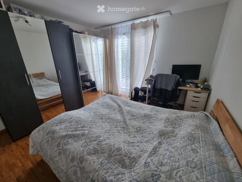 A furnished bedroom with a bed, a desk, a chair, a wardrobe, and a mirror. The room has wooden flooring and large windows with curtains.