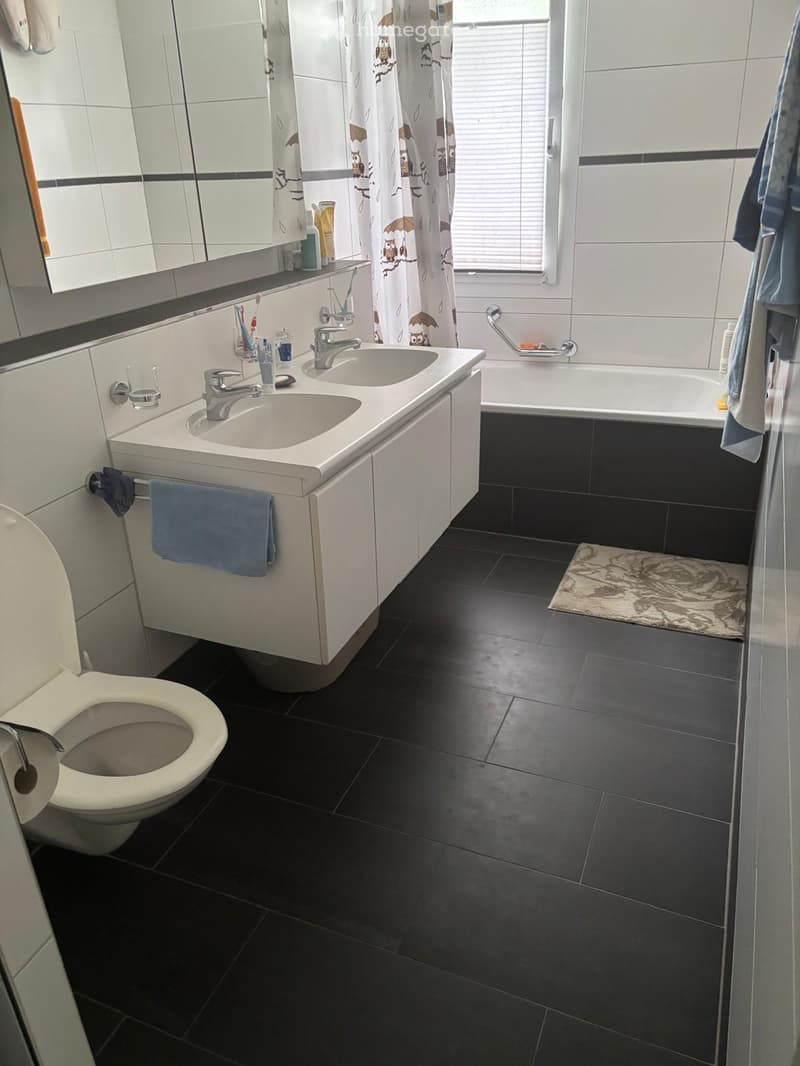 Modern bathroom with black tiled floor, white sink with dual faucets, toilet, bathtub, mirror, and towels.